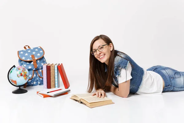 Young joyful attractive woman student in denim clothes, glasses reading book lying near globe, backpack, school books isolated on white background. Education in high school university college concept