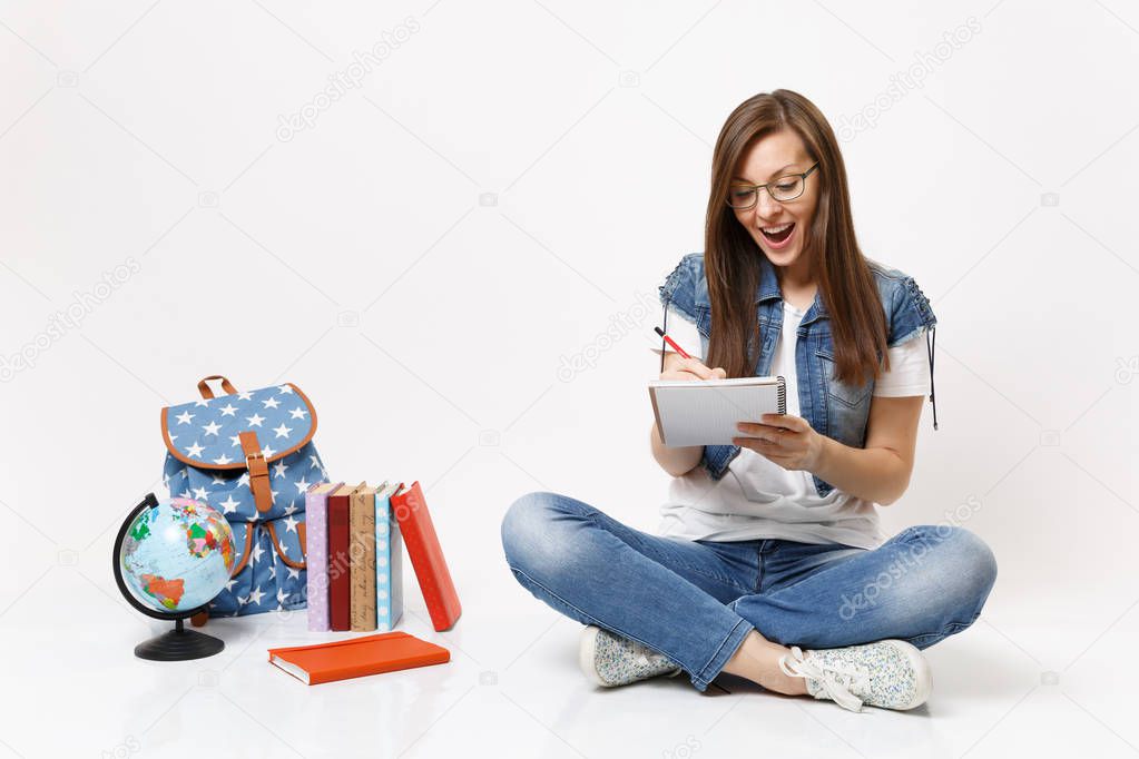 Young happy surprised woman student in glasses writing notes on notebook sitting near globe, backpack, school books isolated on white background. Education in high school university college concept