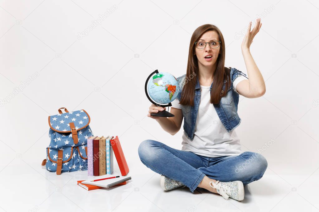 Young puzzled dissatisfied woman student in glasses holding world globe spreading hands sitting near backpack, school books isolated on white background. Education in high school university college
