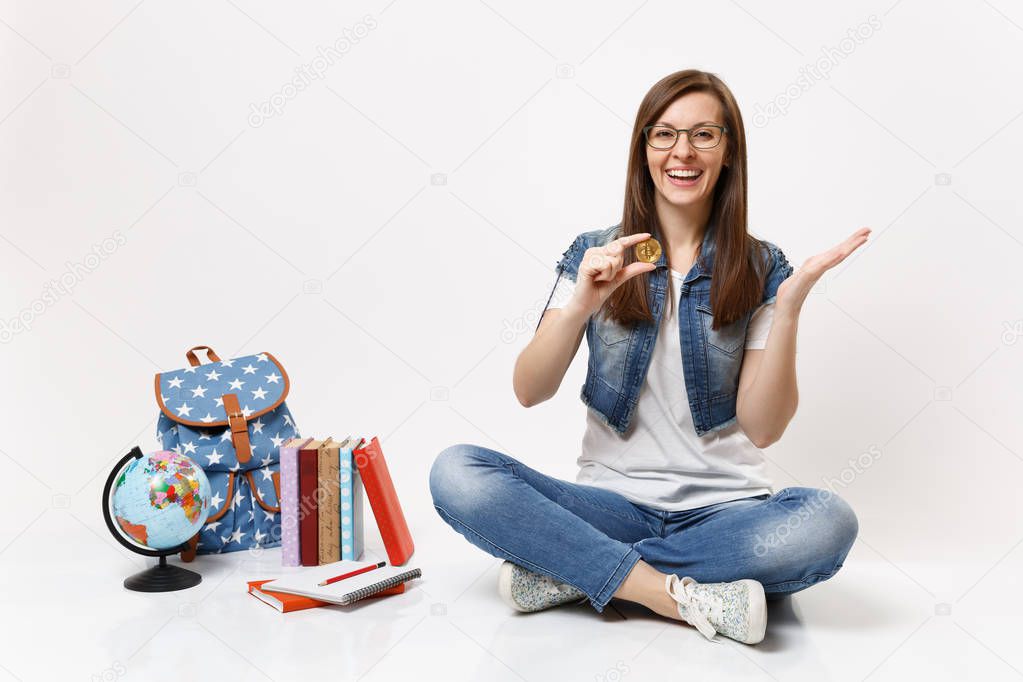 Young laughing happy woman student in glasses holding bitcoin spreading hands sit near globe, backpack, school books isolated on white background. Education in high school university college concept