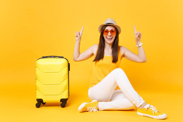 Traveler tourist woman in summer casual clothes, hat sit with suitcase isolated on yellow orange background. Female passenger traveling abroad to travel on weekends getaway. Air journey concept