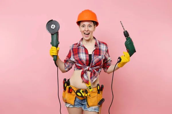 Energy fun handyman woman in gloves, protective orange helmet, kit tools belt full of instruments holding power saw electric drill isolated on pink background. Female in male work. Renovation concept