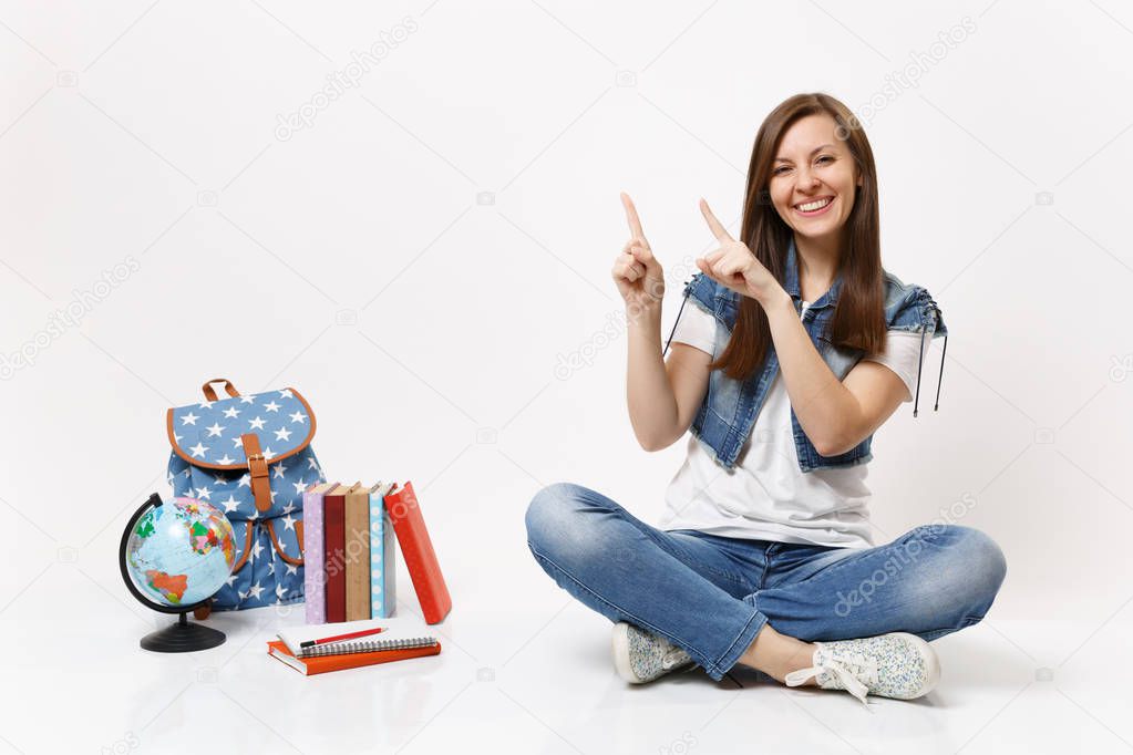 Portrait of cheerful woman student in denim clothes pointing index fingers up aside sitting near globe backpack, school books isolated on white background. Education in high school university college