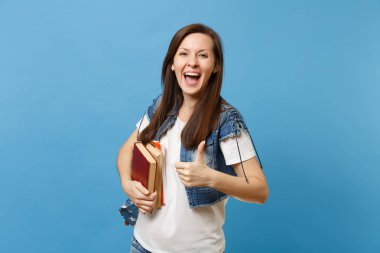 Portrait of young overjoyed woman student with opened mouth with backpack showing thumb up and holding school books isolated on blue background. Education in high school university college concept clipart