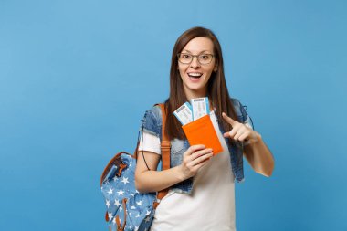 Genç kadın öğrenci bardaklarda sırt çantası işaret parmağı passport boarding pass biletleri üzerinde mavi arka plan izole holding ile hayran. Yurt dışındaki üniversiteler eğitim. Hava seyahat uçuş