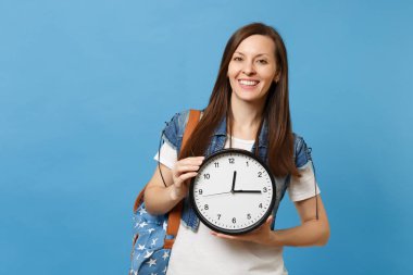 Portrait of young casual smiling attractive woman student in t-shirt, denim clothes with backpack hold alarm clock isolated on blue background. Education in high school. Copy space for advertisement clipart