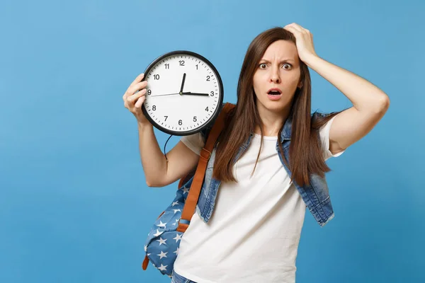 Portrait of young shocked irritated woman student with backpack clinging to head, hold alarm clock isolated on blue background. Time is running out. Education in college. Copy space for advertisement