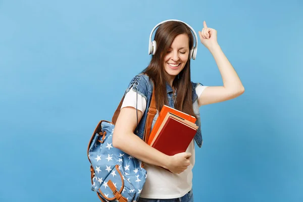 Young cute woman student in denim clothes with backpack headphones listen music hold school book pointing index finger dancing isolated on blue background. Education in high school university college