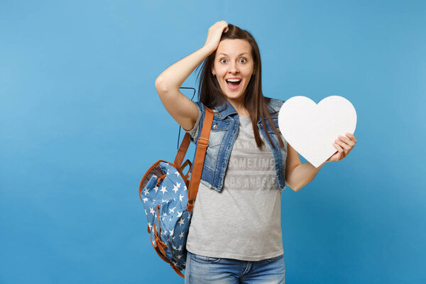 Portrait of young shocked excited woman student with backpack holding white heart with copy space and clinging to head isolated on blue background. Education in college. Copy space for advertisement