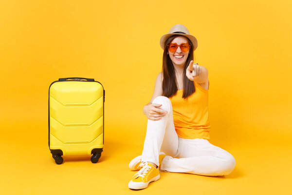 Tourist woman in summer casual clothes, hat sit with suitcase, showing with hand something isolated on yellow orange background. Girl traveling abroad travel on weekends getaway. Air flight concept