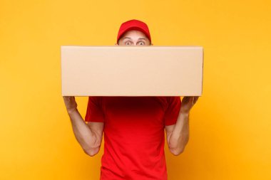 Teslimatçı sarı turuncu arka plan üzerinde izole kırmızı üniforma. Profesyonel erkek çalışan kurye veya Bayi kap t-shirt boş karton kutu tutarak. Paket hizmet anlayışı alma.