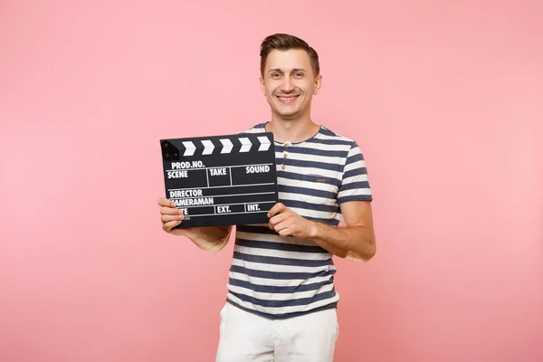 Portrait of young man wearing striped t-shirt holding classic black film making clapperboard isolated on trending pastel pink background. People sincere emotions lifestyle concept. Advertising area