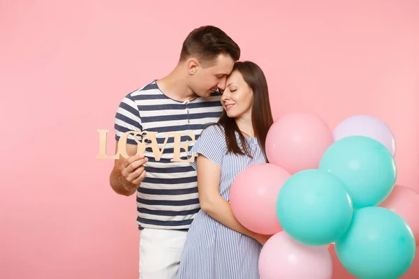 Tender couple hold wooden word letters love. Woman and man in blue clothes celebrating birthday holiday party on pastel pink background with colorful air balloons. People sincere emotions concept