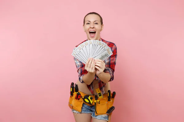 Young handyman woman in plaid shirt, denim shorts, kit tools belt full of variety instruments hold lots of cash dollar money isolated on pink background. Female doing male work. Renovation concept