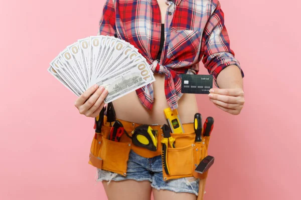 Handyman woman in plaid shirt denim shorts, kit tools belt full of variety instruments hold lots of cash dollar money, credit card isolated on pink background. Female in male work. Renovation concept
