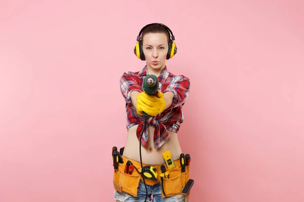 Energy sexy handyman woman in gloves, noise insulated headphones, kit tools belt full of instruments holding power electric drill isolated on pink background. Female in male work. Renovation concept