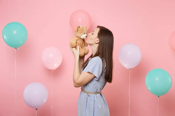 Portrait of attractive young woman wearing blue dress holding and kissing teddy bear plush toy on pink background with colorful air balloons. Birthday holiday party, people sincere emotions concept