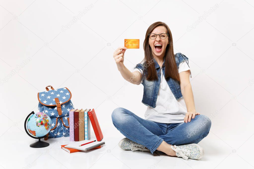 Young crazy overjoyed woman student in glasses screaming holding credit card sitting near globe backpack school books isolated on white background. Education in high school university college concept