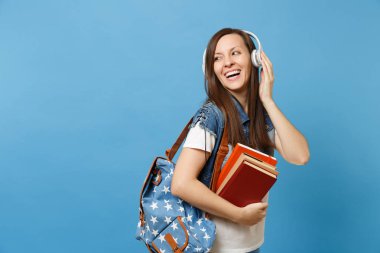 Denim giysileri üzerinde mavi arka plan izole okul kitapları tutan müzik dinleme sırt çantası kulaklık ile genç çok gülen kadın öğrenci portresi. Eğitimde yüksek okul üniversiteler