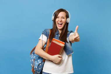Sırt çantası ile denim giysili genç çok memnun kadın öğrenci, okul holding müzik dinleme gösteren başparmak kitaplar kadar kulaklık üzerinde mavi arka plan izole. Eğitimde yüksek okul üniversiteler