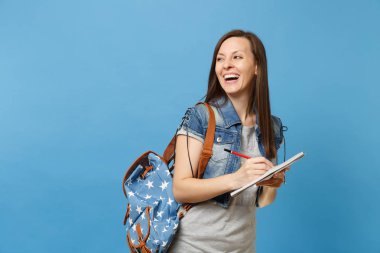 Denim giysiler bir kenara not defter üzerinde yazma kopya alanı üzerinde seyir sırt çantası ile gülen genç kadın öğrenci portresi üzerinde mavi arka plan izole. Eğitimde yüksek okul üniversiteler