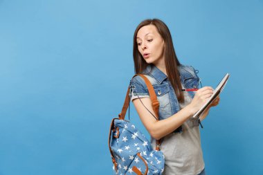 Genç kurnaz meraklı kadın öğrenci sınav gözetleme, üzerinde mavi arka plan izole defter kalem tutan hile alarak sırt çantası ile denim giysileri. Lise Üniversite kavramı kolej eğitim