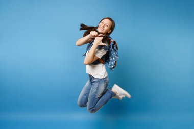 Yan görünümü tam uzunlukta denim giysileri gösteren başparmak yukarıya üstünde izole mavi arka plan atlama sırt çantası ile gülen genç kadın öğrenci portresi. Üniversitede eğitim. Kopya alanı reklam için