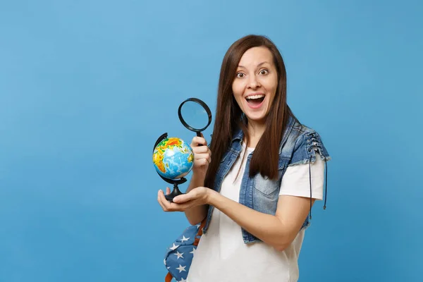 Young excited woman student in denim clothes with backpack looking on world globe with magnifying glass learn about countries isolated on blue background. Education in high school university college