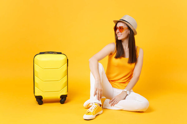 Happy tourist woman in summer casual clothes, hat sit with suitcase, looking aside isolated on yellow orange background. Girl traveling abroad to travel on weekends getaway. Air flight concept