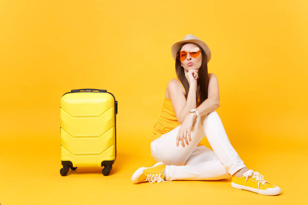Traveler tourist woman in summer casual clothes, hat sit with suitcase isolated on yellow orange background. Female passenger traveling abroad to travel on weekends getaway. Air journey concept