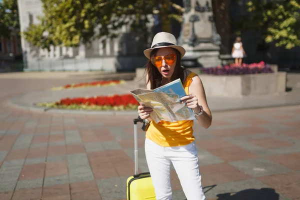 Shocked puzzled traveler tourist woman in orange heart glasses with suitcase search route on city map in city outdoor. Girl traveling abroad to travel on weekends getaway. Tourism journey lifestyle