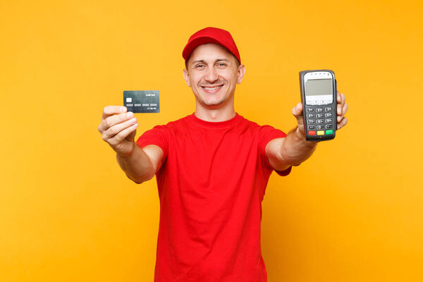 Delivery man in red uniform isolated on yellow background. Male employee in cap, t-shirt courier holding wireless modern bank payment terminal to process and acquire credit card payments, black card
