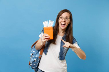 Genç kadın öğrenci bardaklarda pasaportla işaret parmağı işaret, pas bilet üzerinde mavi arka plan izole yatılı sırt çantası ile şaşırttı. Yurt dışındaki üniversiteler eğitim. Hava seyahat uçuş