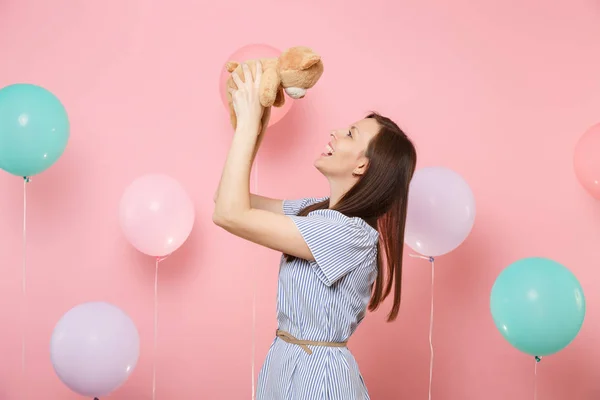 Portrait of laughing pretty happy young woman in blue dress holding teddy bear plush toy on pastel pink background with colorful air balloons. Birthday holiday party, people sincere emotions concept