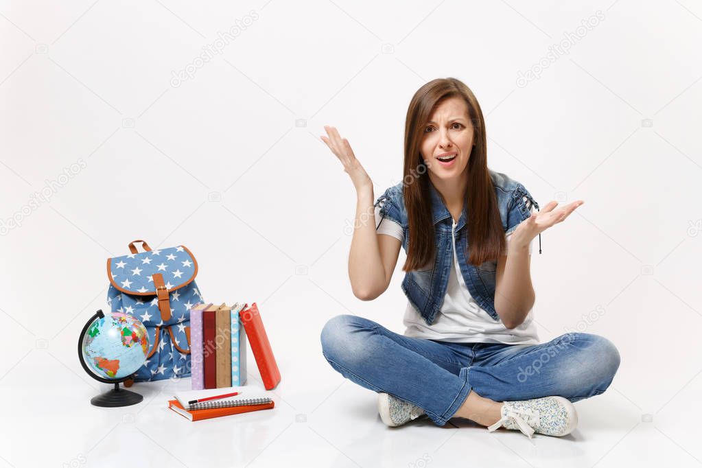 Portrait of young concerned irritated woman student in denim clothes spreading hands sitting near globe backpack school books isolated on white background. Education in high school university college