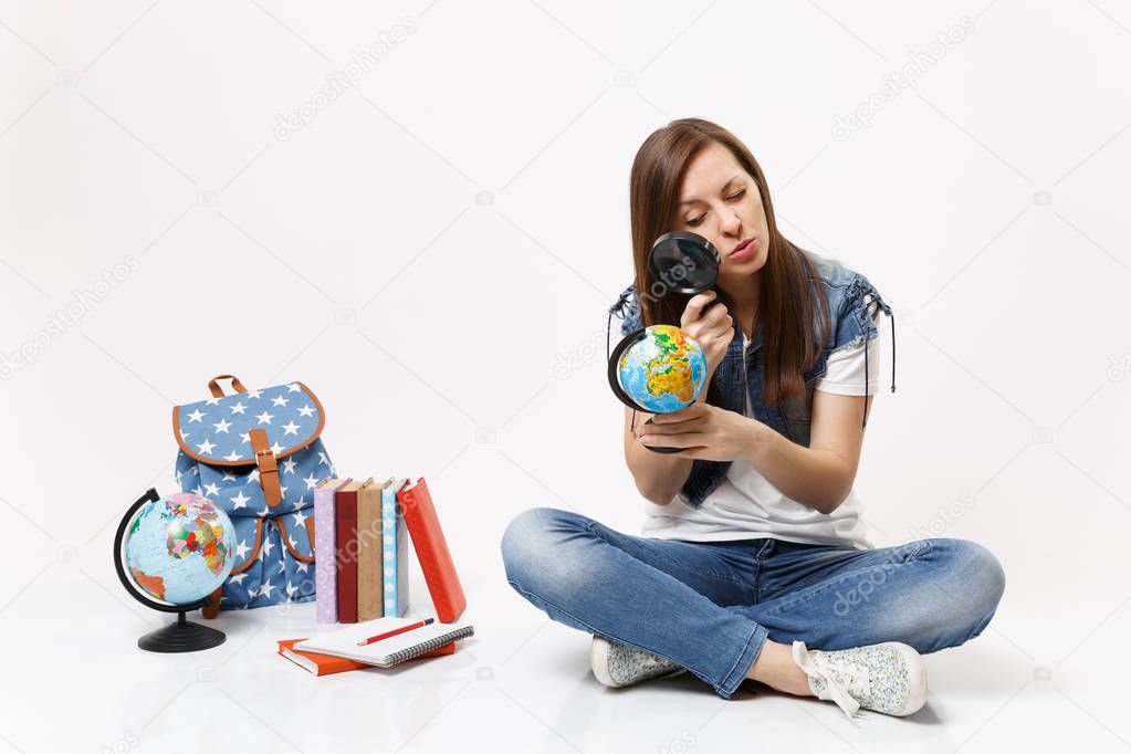 Young smart interested woman student looking on globe with magnifying glass learning sitting near backpack, school books isolated on white background. Education in high school university college