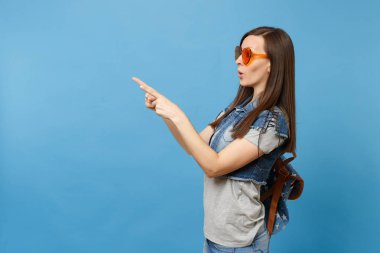 Yan görünümü genç kadın öğrenci sırt çantası turuncu kalp gözlük dizin üzerinde mavi arka plan izole parmak işaret içinde ile açılan ağzı ile şok. Yüksek okulda eğitim. Kopya alanı reklam için