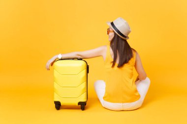 Arkadan görünüm turist kadın yaz rahat kıyafetler şapkalı oturmak sarı turuncu arka plan üzerinde izole çanta. Hafta sonu kaçış üzerinde seyahat için yurt kadın yolcu. Hava uçuş kavramı