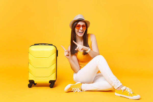 Traveler tourist woman in summer casual clothes, hat sit with suitcase isolated on yellow orange background. Female passenger traveling abroad to travel on weekends getaway. Air journey concept