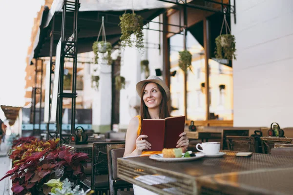 Woman in outdoors street coffee shop cafe sitting at table in hat, reading book with cup of cappuccino, cake, relaxing in restaurant during free time. Mobile Office in summer. Lifestyle rest concept