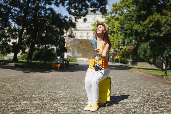 Happy traveler tourist woman in yellow clothes, hat sitting on suitcase holding city map search route in city outdoor. Girl traveling abroad to travel on weekends getaway. Tourism journey lifestyle