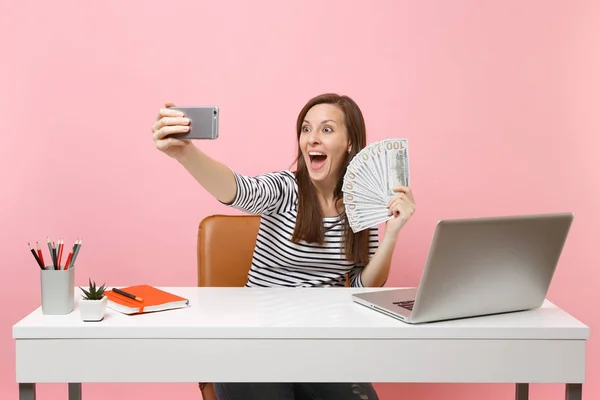 驚く女性撮影 Selfie ドルの現金お金のバンドル多くを押しながらピンク色の背景に分離されたノート パソコンのデスクに座って仕事の携帯電話で撮影を行います ビジネス キャリアを達成 コピー スペース — ストック写真
