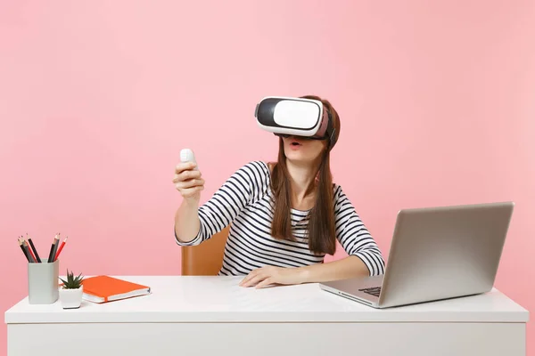 Young amazed woman in headset of virtual reality on head using remote control sit work at white desk with pc laptop isolated on pastel pink background. Achievement business career concept. Copy space