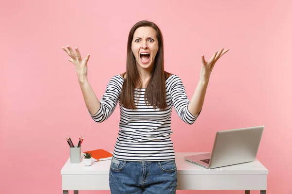 Young angry woman screaming and spreading hands work standing near white desk with pc laptop isolated on pastel pink background. Achievement business career concept. Copy space for advertisement
