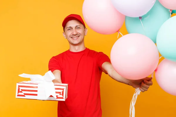 Delivery man in red uniform isolated on yellow orange background. Male employee in cap, t-shirt courier dealer holding bunch of colorful pastel air balloons, red gift box. Service concept. Copy space