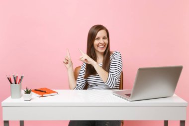 Genç gülmek kadın rahat kıyafetler noktası işaret parmağı bir kenara oturmak, pastel pembe arka plan üzerinde izole çağdaş pc dizüstü bilgisayar ile beyaz masa başında çalışmak. Başarı iş kariyer kavramı. Kopya alanı