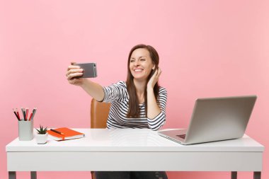 Sit pastel pembe arka plan üzerinde izole çağdaş pc dizüstü bilgisayar ile beyaz masa başında çalışma sırasında cep telefonunda alarak selfie yapıyor neşeli kadını vurdu. Başarı iş kariyer kavramı. Kopya alanı