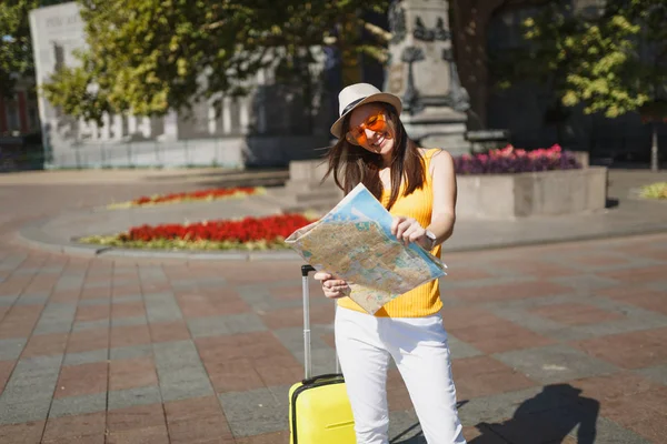 Laughing traveler tourist woman in hat, yellow clothes, orange heart glasses with suitcase hold city map in city outdoor. Girl traveling abroad to travel on weekend getaway. Tourism journey lifestyle