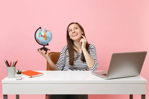 Young pensive woman holding globe, dreaming planning vacation while sit, work at office with contemporary pc laptop isolated on pastel pink background. Achievement business career concept. Copy space