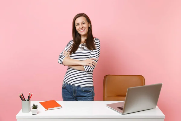 Young successful girl in casual clothes work standing near white desk with contemporary pc laptop isolated on pastel pink background. Achievement business career concept. Copy space for advertisement